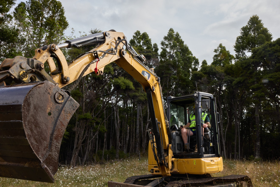 Man operating excavator in a field with Kauri forest in the background.